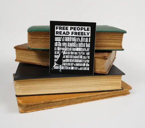 A rectangular sticker made from a linocut print of a person reaching for a book on a large bookshelf, with “free people read freely” above it