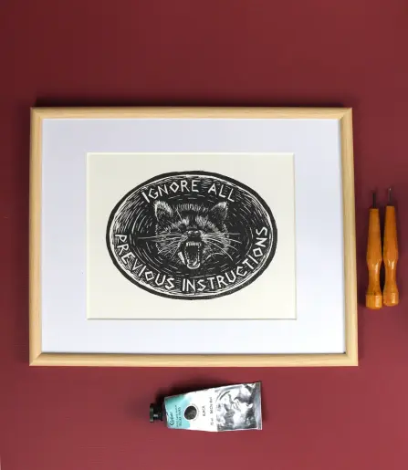 A linocut print featuring an oval with a raccoon in the middle, and “ignore all previous instructions” around the border