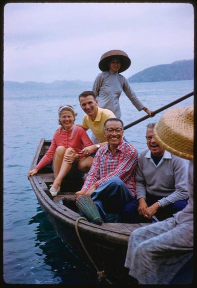 Image of a group of people sitting in a boat on water, with mountains visible in the background and one person standing at the back holding onto a pole. The woman seated second from left is wearing sunglasses and smiling while looking directly into the camera. Next to her is another man who appears cheerful as he looks over his shoulder towards others in the group. Behind him sits an older gentleman leaning forward with glasses on, also beaming for the photo. To his right is a younger individual sitting down, seemingly relaxed but not engaging much with the surrounding people or looking at the camera directly. Another woman to the far left of this man seems calm and smiles slightly towards the viewer.
In front of all these individuals are two women; one wearing pink attire sits on the edge of the boat holding onto its side while another is in grey, sitting down but not interacting with others around her. A straw hat partially obscures part of a person's head at the bottom right corner, and an elderly man is visible to the far left outside of this group, only showing his shoulder area.
The overall mood appears jovial as everyone looks content or happy during their time together on water.