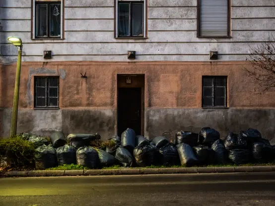 Teil einer Fassade eines städtischen Wohnhauses, das schon bessere Zeiten gesehen hat.
Der Verputz ist unten rostrot, die Etage darüber beige.
Auf dem Grünstreifen vor dem Haus türmen sich schwarze Müllsäcke.