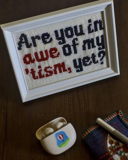 a cross stitch framed in a white frame that reads

are you in
awe of my
'tism yet? 

there's wireless earbuds, a blue notebook with a fountain pen above it. 

these are placed on a wooden table.