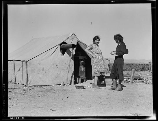 The image depicts a scene from the past, captured in black and white. It features two women standing outside of a makeshift tent structure with fabric walls supported by poles on an unpaved ground. One woman is seated on what appears to be a wooden stool or low step, wearing a dress patterned with vertical lines and dark-colored skirt, while engaging in conversation with the other woman who stands beside her.

The second woman holds a notebook or clipboard and pen in hand as she writes down notes, suggesting that she might be conducting an interview or survey. She is dressed more formally than the seated woman; wearing a long-sleeved dress paired with dark-colored trousers, shoes, and gloves. Her posture indicates attentiveness to her task.

The environment around them reveals signs of rural life, possibly in a farming area as suggested by the context provided below the image: "Near Calipatria, Imperial Valley, California. In grower's camp for migrant labor on the edge of the pea fields. Public health nurse interviews and advises prospective mother, aged seventeen, before arrival of first baby. Husband, aged twenty-three, is out picking. Made seventy-three cents this morning." The mention of a public health nurse implies that they may be partaking in some form of social service or welfare program.

The overall composition suggests the image captures an interaction between [...]