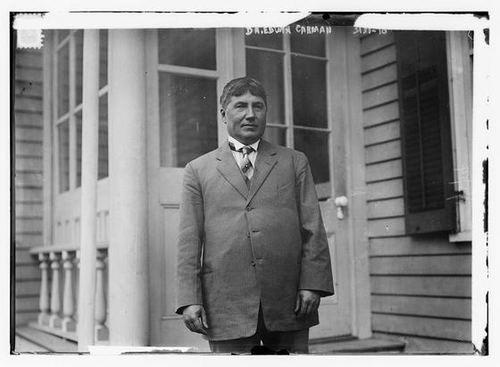 The image is a black-and-white photograph of a man standing in front of a building. The man is dressed in formal attire, wearing a suit with a tie. His posture is upright, and he appears to be facing the camera directly. The building behind him has a classic architectural style, with large windows and a door. The windows have a grid-like pattern, and the door is framed with a simple design. The man's name, "Dr. Edwin Carman," is written in cursive at the top of the image, and there is a date or number, "5130-18," written in smaller text to the right. The overall tone of the image suggests it is from an earlier time period, possibly the early 20th century.