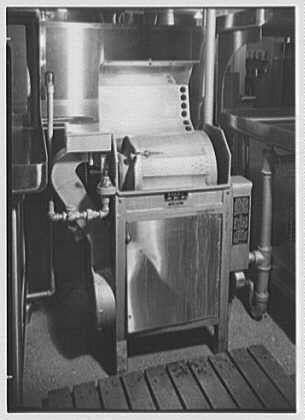 The image depicts a vintage, industrial-grade dishwasher or washing machine from the year 1948. It's made of metal with a large rectangular body and various control knobs on its front panel. The device has visible piping for water supply and drainage attached to it. The setting appears to be within an industrial kitchen environment as suggested by surrounding appliances in the background, possibly indicative of a restaurant or food service establishment during that period.

The black-and-white photograph captures the machine's metallic surfaces with reflections from overhead lighting, giving the image a somewhat aged look typical for documents and photographs from the mid-20th century. Notably, this particular photo is credited to Gottschalch-Schleisner Inc., which suggests it may have been part of an archive or collection related to commercial photography during that era.

Additional context includes references like "Schrafft's" and "Esso Building," Rockefeller Center in New York City, indicating the image could be connected with a historical account about these locations. It is also mentioned as having one negative measuring 5x7 inches, which gives insight into its original dimensions when developed for printing purposes.

The presence of an alt text helps provide context to visually impaired users by describing what they might see if viewing this photograph directly; i [...]