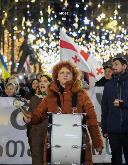 In front of protesters crowd a woman hits into large drum to bring a rhythm and energy into crowd walking. Some people in crowd hold large signs and flags.
New year decorations visible at top, out of focus so they blurred.