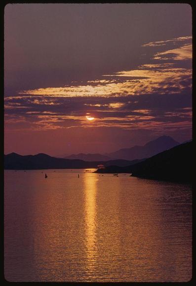 This image captures a breathtaking sunset over the waters near Hong Kong during the summer of 1959. The warm hues of orange and pink paint the sky as the sun dips below the horizon, reflecting off the still water's surface. Silhouetted mountains rise in the distance under this vibrant backdrop, while small boats dot the expanse of water, adding life to the tranquil scene.

Incorporating elements like a parachute flying above the calm waters and sailboats scattered across the landscape suggest an air of leisurely exploration typical during that era's travel. The photograph is credited as part of Sports Illustrated coverage by renowned photographer Toni Frissell, known for her pioneering work in various fields including sports, fashion, and travel photography.

A black and white variant exists alongside this color transparency image, showcasing a broader range of tones from the vibrant sunset to the subdued shadowed areas near Hong Kong. This dual-tonal approach provides an intriguing contrast between colors and grayscale nuances that emphasize different aspects within the scene captured by Frissell in her 1959 travels through X5826's lens.

Toni Frissell was a pioneering figure in photography, particularly for women photographers of her time who broke barriers to gain recognition. Her distinctive style often featured unique perspectives and compositions, capturing not just [...]