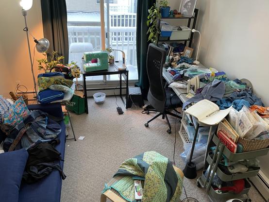 A messy apartment living room with a mint-green sewing machine in the balcony window. A long cutting/pressing table is heaped with projects.