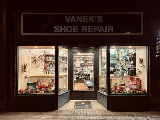 Storefront for Vanek’s Shoe Repair. The inside is lit up brightly in contrast with the dark night. Inside are lots of boots and shoes on shelves with repair tags hanging from them, and lots of various repair machines and tools.