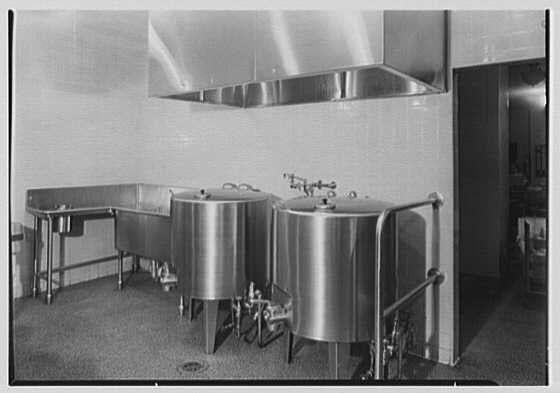 The image depicts a vintage, monochromatic photograph of an industrial kitchen or brewing facility. It shows several large stainless steel vessels lined up against the wall with various pipes and fittings attached to them. The containers are likely used for fermentation processes in food production, such as beer making.

Above these vessels is what appears to be a metal exhaust hood designed to capture odors and heat generated during cooking or processing. On the left side of the image stands a rectangular stainless steel table or prep area with several smaller items on top that seem out of focus due to their distance from the camera lens.

The setting has an industrial, utilitarian feel with bare walls, visible pipes running along them, and a nondescript floor. The overall ambiance suggests this photograph might be documenting early 20th-century commercial food preparation methods or brewing techniques used during that time period in New York City's Rockefeller Center complex as mentioned in the caption.

The black-and-white nature of the image adds to its vintage appeal, providing an insight into how such facilities were constructed and operated more than seven decades ago.