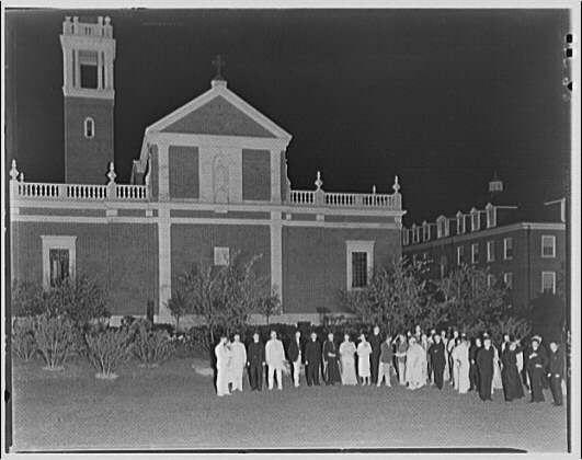 A black and white photograph showing a group of people standing outside at night, with the Bishop's residence in the background. The individuals are dressed formally; most wear long coats or dresses while some have hats on their heads. A few women can be seen wearing headscarves or veils over their hair. They stand organized into three rows - two lines and one line of people behind them, all facing towards the camera with a slight smile. Behind this group is an elegant brick building featuring large windows and white decorative elements above its entrance which bears religious symbolism such as crosses atop towers in what appears to be church architecture.