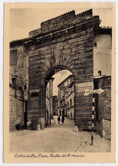 This black and white photograph depicts a historic stone archway known as Porta di S. Maria in Città della Pieve, Italy. The architecture of the arch is robust with visible texture from weathering over time. Beyond the entrance, an alleyway unfolds leading to several brick buildings that line either side of the path. A few individuals are seen walking along this narrow street, providing a sense of scale and life within what appears to be an old town setting.

On the right-hand wall outside the archway is a sign with "VEICOLI" pointing towards vehicles, suggesting it's likely a one-way or traffic-controlled passage. The overall scene conveys a quiet moment in time, possibly from several decades ago given the style of the photograph and attire of the people visible within this vintage image.

The condition of the paper suggests age; there are slight creases and some discoloration around the edges, indicating that it is not just an old print but also itself has been through a period. The bottom left corner bears handwritten text in cursive script: "Città della Pieve. Siove. Porta di S. Maria," which translates to "City of Peace" and likely identifies this landmark within the town.

This photograph provides both historical context as well as artistic value, capturing not only a physical structure but also an aspect of daily life in Città della Pieve during its time period.