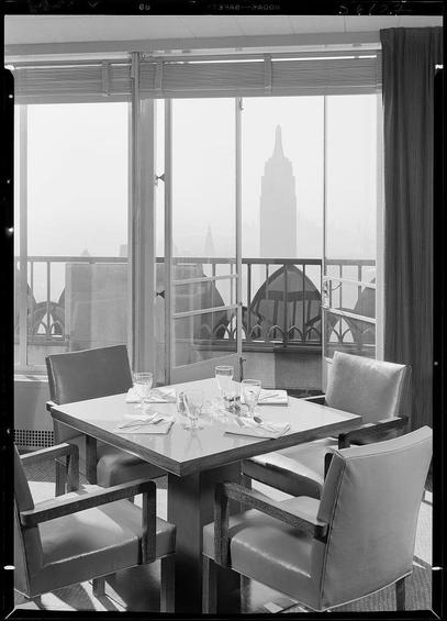 The image depicts a black and white photograph of an elegantly set dining table on what appears to be the terrace or balcony of a high-rise building. The view from this vantage point showcases several iconic structures, including the Empire State Building in the distance, indicating that the scene is likely located within New York City.

In the foreground, we see a rectangular wooden table surrounded by six chairs with leather upholstery and metal accents. On the tabletop, there are neatly arranged place settings for four individuals, featuring glassware, silverware, folded napkins, and plates suggestive of an upcoming formal meal or gathering.

The backdrop reveals large windows that frame expansive views beyond the cityscape, adding a sense of openness to the indoor setting. The overall atmosphere exudes sophistication and exclusivity associated with high-end dining establishments in New York City's Rockefeller Center vicinity during 1949.