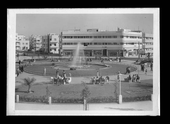 The image shows a historical black and white photograph of Dizengoff Circle in Tel Aviv, captured from the north. The view depicts an urban square with modernist architecture dominating the background. Several mid-20th-century buildings surround a central fountain where water is seen spraying upwards, creating a focal point for people gathered around it.
People are scattered throughout the plaza; some sit on benches while others stroll or stand in groups engaged in conversation. The foreground features manicured lawns and pathways enclosed by metal railings, with palm trees dotting the landscape. A few individuals can be identified among the crowd as they appear relatively small compared to the overall scene.
The image is credited to Matson Photo Service and seems to have been taken between 1934 and 1939 from a negative made during that period (between 1934 and 1946). This indicates its vintage nature, reflecting Tel Aviv's development in those years.
