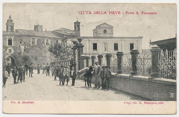 The image is a black and white postcard depicting an outdoor scene of the historical town gate known as Porta S. Francesco in Città della Pieve, Italy. The photograph captures multiple individuals from different age groups walking along what appears to be a paved street adjacent to ornate metal gates leading into or out of a walled area with classical architecture visible behind them.

In the foreground, there are at least 12 people scattered across the frame: some standing, others in motion. Among these figures, adults and children can be distinguished by their attire—a mix of casual clothing typical for the period, including hats and coats for men, dresses or skirts for women, and what appears to be a child's school uniform.

The architecture includes buildings with stone facades, large windows, decorative elements such as pilasters on columns, railings along pathways, and ornate gates. Notably, there is an arched gateway leading into the walled area which has metal grills or fencing for security purposes.

In the background stands a church tower topped with what appears to be a cross, indicating its religious function within the community. The sky above seems overcast, casting soft light on the scene without harsh shadows. There are no visible modern elements in this historical snapshot; it maintains an authentic representation of past times when Città della Pieve was  [...]