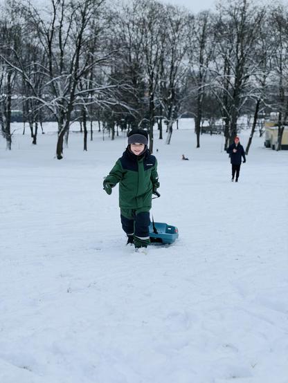 Young child wearing moss green and navy blue winter jacket with black hat, smiling while pulling ocean green plastic sled through snow-covered park. Snow-laden bare deciduous trees fill background. Child’s father visible in distance. Fresh snow covers ground with visible sled tracks. Overcast winter morning in Grīziņkalns park, Rīga. Child appears mid-walk, actively sledding.