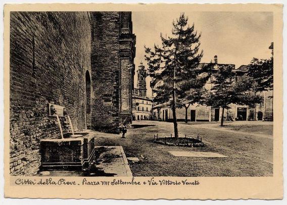This black and white photograph captures a historical European street scene, likely from the early 20th century. The image depicts an old building with stone walls on the left side that appears to be part of a church or cathedral, given its architectural style which includes arched windows and what seems like religious iconography above them.

On the right, there is another large structure with multiple levels, including what could be living quarters for clergy members. Between these buildings are trees and greenery, suggesting an urban environment that still has some natural elements within it.

The street itself looks worn down and made of cobblestones or a similar material typical in older towns. There's a fountain or water pump on the left with writing above it which is not legible from this angle but possibly indicates its purpose or donor information, as such features were often commemorated by individuals or institutions during that era.

A figure can be seen walking along the street towards what appears to be an arched entranceway leading into a courtyard. The overall atmosphere of the photograph conveys a sense of history and tranquility, with no modern elements in sight, reinforcing its historical context.

The image is marked at the bottom left corner with handwritten text "Città della Pieve - Piazza VIII Settembre e Via Vittorio Veneto," indicating that this l [...]