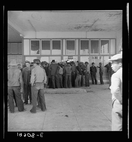 A black and white photograph shows a group of men standing in line outside a building. The individuals are primarily dressed in work attire, with many wearing hats or caps. Some have on jackets while others wear shirts without sleeves; all appear to be adults. They stand shoulder-to-shoulder along the edge of what seems to be an open plaza or courtyard area. In the background is a large structure that could possibly serve as a government building given its formal architecture and multiple windows with visible signs above them, although it's unclear from this perspective what those signage indicates.

There’s text written on the image in white lettering at various positions: "1028- -E" positioned near the bottom left corner; there are two other instances of obscured numbers or letters just below where a timestamp would typically be located. The photograph gives off an atmosphere suggesting it could have been taken during mid-century, likely reflecting social and economic conditions typical in that era.

The image has visible signs of wear and age like fading which might imply historical context to the moment captured as well. Notably, no specific individuals are identifiable or focused on individually; instead, they collectively represent a group from an unspecified socio-economic background possibly related to agricultural labor during harvest season given photographic ca [...]