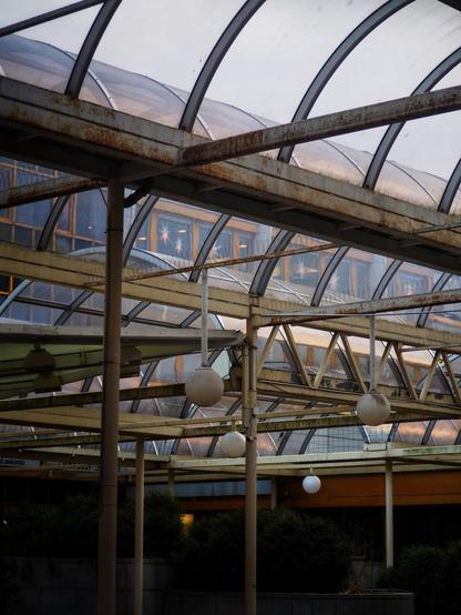 An indoor area featuring a combination of metal and glass structures. The ceiling is arched with visible rust, and there are round light fixtures hanging from the framework. The background shows large windows with reflections, and some greenery is present at the base.