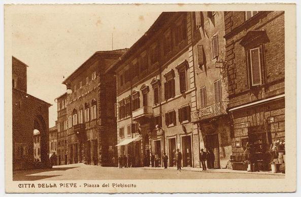 The image is a sepia-toned vintage photograph depicting an old European street scene, possibly from the early 20th century. The architecture suggests it's set in Italy or another Mediterranean region with historic buildings lining both sides of the road.

On the left side, there is a large archway leading into what appears to be a market area or alleyway, while on the right, various shops and cafes are visible under signage such as "Caffè" indicating coffee houses. People dressed in early 20th-century attire can be seen strolling along the sidewalks, some walking alone, others in pairs.

Prominent buildings include one with large windows above a covered entrance that reads "PRAFFANTINI," suggesting it may have been used for social gatherings or events. Another building to its right has shutters and a signboard indicating "RESTAURANTE" which translates from Italian as restaurant. The street is paved, and the absence of modern vehicles suggests transportation methods were more pedestrian-friendly at this time.

The photograph carries text in what appears to be German below it stating: "CITTÀ DELLA PIEVE - Plazza del Plebiscito," indicating that the location captured by the image is a square called Piazza del Plebiscito. The overall condition of the photo indicates wear and age, with some discoloration visible on its surface.

The landmark associated with this photograph can [...]