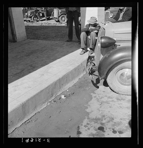 The image is a black and white photograph capturing a moment in what appears to be the early-to-mid 1900s, possibly during the Great Depression era. It depicts several individuals around vehicles on an urban street or roadside setting. One central figure sits alone against the curb of a paved surface, with their legs hanging down beside a vintage car's fender and tire. This person wears casual attire including jeans and shoes, has dark hair peeking out from under a hat, and appears to be resting or waiting.

Another individual is partially visible on the right side inside what looks like an older model automobile, suggesting they are either sitting in the driver’s seat or passenger's area of this vehicle. Their attention seems directed towards something outside the frame of the photo.

In the background, there are two people standing near another vintage car with a metal container attached to its back; one person is bending over as if interacting with the object on top of it and appears focused on their task. The other individual stands beside them, dressed in work clothes including what looks like denim pants.

The surroundings include cracked pavement or curb edges, indicative of wear from usage or age, alongside a larger vehicle parked further back which could be related to farm equipment due to its size and design features such as a large front grille.

Overall, the s [...]