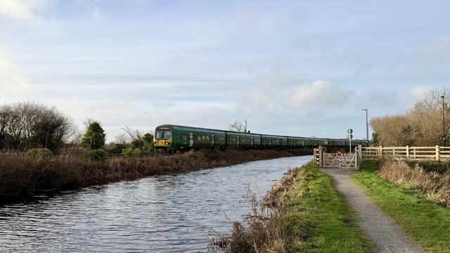 A wide-screen landscape shot looking down a gravel trail along the left bank of a canal with a railway line on the right bank and a long green railcar-type train with a distinctly square shape and a yellow front. The sky above is a hazy blue, and the water is looking a little choppy.