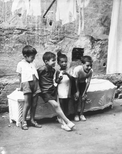 The image is a black and white photograph depicting four children outdoors. They are gathered around an ornate, vintage-style coffin that has been repurposed as seating or possibly for playing on. The environment appears to be somewhat rural with laundry hanging from above, indicating it might be in front of a home.

From left to right: the first child is wearing a light-colored shirt and dark shorts, standing upright next to the box; the second child sits inside the coffin-like structure, leaning back comfortably; the third stands on his toes outside the coffin, slightly hunched over with hands clasped together at chest level as if in conversation or playfulness. The fourth child mirrors the first's posture but is more fully engaged with a piece of paper or card.

The background includes walls made from rough-hewn stones and other indistinct items suggesting an outdoor domestic setting that might be indicative of economic hardship, given the utilitarian repurposing of the coffin for such casual activities. The overall mood seems to capture moments of childhood playfulness amid modest surroundings.