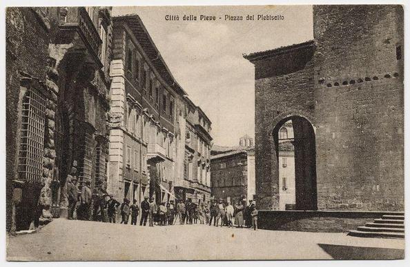 The image is a black and white vintage photograph depicting an urban street scene. It appears to be from the early 20th century, given its monochromatic nature and style of dress worn by people in the photo.

In this setting, we see several individuals standing on what seems like a paved walkway or square within a historical cityscape. The architecture is distinctly European with buildings exhibiting classical facades featuring columns, pilasters, and arched windows. One prominent structure to the right has an arch leading into it which might be indicative of some public building.

The people are dressed in attire typical for that era: men wearing hats such as bowlers or caps, while women wear dresses with skirts reaching mid-calf; there is no visible use of modern clothing like t-shirts and jeans. The crowd appears to be gathered around a central event or attraction not fully captured within the photo's frame.

The photograph has inscriptions at the top that read "Città della Pieve - Piazza del Plebiscito," suggesting this scene takes place in Piazza del Plebiscito, which is likely located in Città della Pieve. The term 'plebiscito' refers to a referendum or plebiscite.

There's also an additional image referenced with the identifier "673316fa89b10e7a26e5135d," which seems unrelated and could be associated with another context, perhaps pertaining to historical records. H [...]