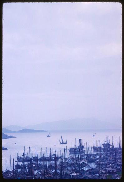 A view of a harbor filled with boats on the water, with mountains in the distance and sailboats sailing. The photograph has an overall blue tint to it. Additional information about this image can be found in Hong Kong - X5826, Extra 1959 from Toni Frissell's collection.