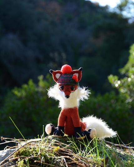 Petit renard au crochet, photographié en extérieur, assis sur un rocher couvert de mousse et d'herbe. Il porte un bonnet vert et rouge avec un pompon rouge et des lunettes rondes. Sa queue blanche est duveteuse. En arrière-plan on distingue une forêt flou.