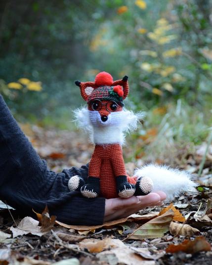 Petit renard au crochet, assis dans une main posée au sol, photographié en extérieur sur un tapis de feuilles mortes. Il porte un bonnet vert et rouge avec un pompon rouges et des lunettes rondes. Il présente une grande queue blanche duveteuse. L'arrière-plan est flou, avec des tons verts et automnaux.
