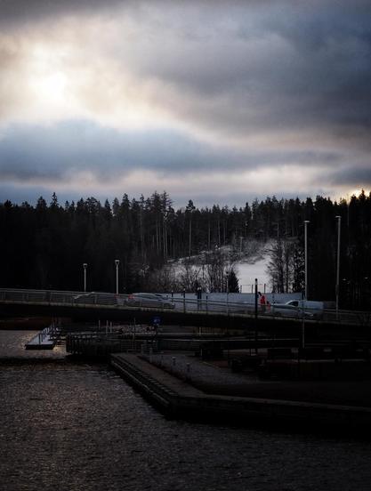 A bridge crosses a body of water, with a few cars on it. In the background, a forest is partially covered in snow under a cloudy sky. The landscape has a cold, wintery atmosphere.