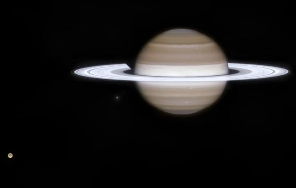 In this JWST image, Saturn appears as a pale gold/brownish sphere with horizontal bands, encircled by a bright whithish, thin oval ring system. The atmosphere is showing off a few circle whithish storm-like features. On the bottom left also its world Titan is visible as a gold glowing sphere.