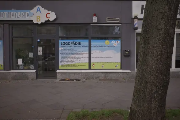 A storefront of a logopedics office, the trunk of a tree is covering up the shopwindow on the right which is from a barber shop.