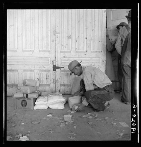 The image depicts a man in his 30s or early 40s crouched down and looking at several bags of flour. He appears to be working on the ground, possibly sorting through broken pieces of paper or debris scattered around him. His attire includes overalls with suspenders, a long-sleeved button-up shirt under an apron-like garment, and a hat that provides some shade from above.

Behind him stands another man in similar clothing, suggesting they are part of a work crew or involved in agricultural labor during the pea harvest season mentioned in the context. The setting is outdoors with wooden doors behind them featuring vertical lines and metal hardware. There's an overall sense of hardship and manual labor as indicated by their posture and environment.

The image captures a historical moment likely from around 1938, given it was taken outside the Farm Security Administration (FSA) grant office during that time period in California when there was significant agricultural work being done despite financial difficulties.