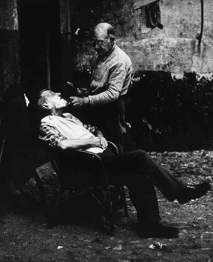 The image depicts a scene of two elderly men engaged in an activity outdoors. One man is seated on what appears to be a chair, with his legs crossed and arms resting along the armrests or supports behind him. His face shows signs of weariness or discomfort as he looks upwards.

The other man stands beside him, leaning over closely enough to engage directly with the first man's mouth area. This standing man is holding an object in his right hand that seems to be a brush or some kind of grooming tool, suggesting that he might be performing a task such as shaving or dental care on the seated individual.

The setting appears casual and unrefined, possibly indicative of a street-side barber shop or similar establishment where services are provided at no cost. The background is dark and indistinct but hints at an outdoor environment with some scattered objects around them.

Both individuals exhibit signs of aging such as wrinkles and balding heads. Their attire consists of simple, casual clothing suitable for everyday activities rather than professional work uniforms.

The photo captures a moment that conveys a sense of intimacy or familiarity between the two men, possibly hinting at an established routine or bond in their interactions with one another despite any apparent discomfort on both parties' faces.