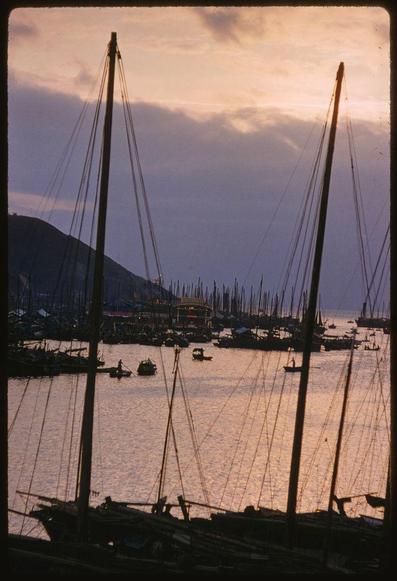 A colorful photograph taken at dusk in a harbor filled with boats and yachts, depicting their sails and masts. In the distance is a hill with lights on top of it while various other vessels dot the water surface which reflects some light possibly from a nearby source or city lights as well. The photo has an old look to it indicating its age could be around 1950s based on information provided in reference link.