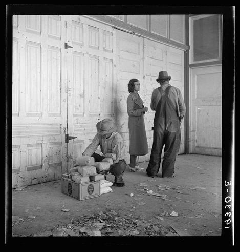 The image is a black and white photograph depicting three individuals in an industrial or agricultural setting. The environment appears to be a warehouse with wooden doors, where the ground shows signs of neglect such as scattered debris.

One person, presumably male based on clothing style and posture, is seated on the concrete floor outside a door, handling what looks like bags filled with items possibly related to farming produce given context clues from the surroundings. He wears overalls, shoes that appear worn out, and has his sleeves rolled up for manual work. His attention seems focused on the task at hand.

Another individual, likely female due to attire, stands beside him wearing a coat and skirt, her hands clasped in front of her as she observes or waits patiently near an open door which appears frequently used given its worn state. The third person is standing further away from them with their back partially turned towards the camera; this figure seems male based on clothing style—a hat and overalls—and his stance suggests he might be taking a break or observing something outside of view.

The overall atmosphere conveyed by the image exudes hardship, possibly during economic struggles reflected through the worn conditions around them. The photograph's context comes from Outside the Farm Security Administration (FSA) grant office during pea harvest in Calipatri [...]