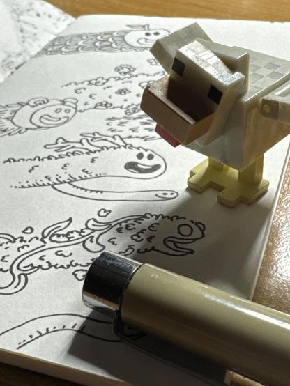 A small toy figure of a Minecraft chicken astands on an open sketchbook featuring playful, hand-drawn illustrations of weird and smiling characters. A pen lies next to the sketchbook