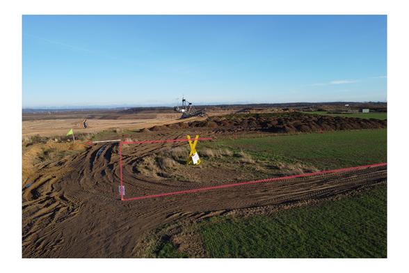Foto einer Tagebau-Kante. In das Bild sind rote Linien eingezeichnet, die ein Grundstück auf einer Wiese markieren. Ein Teil der Wiese innerhalb der Markierung ist bereits abgebaggert. Im Hintergrund ein Schaufelradbagger."