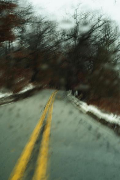 Color photo taken out a wet windshield, looking up a two lane highway with snow on either shoulder and a clump of gnarly leaf-less trees in the midground, as the road disappears over a. little ridge.