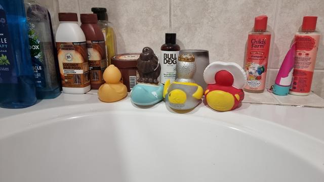Four thematic rubber ducks sit along the edge of a fitted white bathtub, seemingly deliberate in their placement which sees them facing directly away from the bather and towards various shampoo bottles and a candle in a glass.