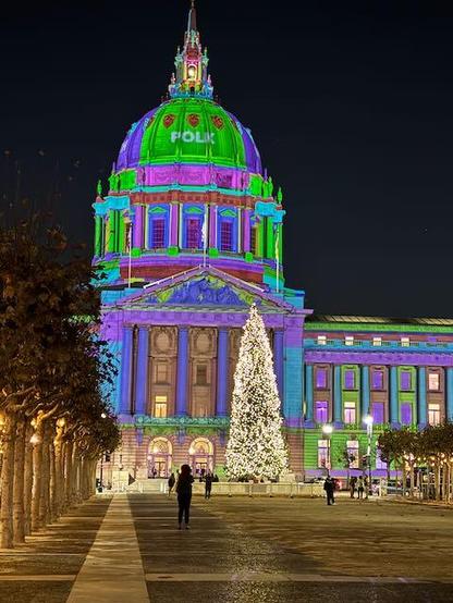 The facade and dome of San Francisco city hall lit in blues, greens and purples, plus a large tree with white lights.