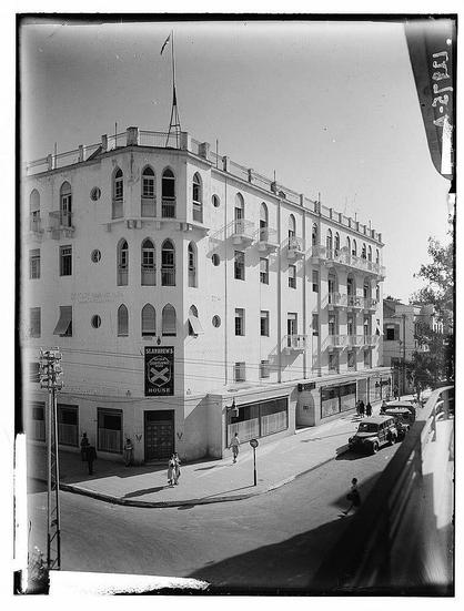 The image depicts a black and white photograph of an urban street scene. It shows the corner view of a multi-story building with architectural features such as arched windows, balconies, and decorative balustrades on its upper floors. The facade has several rectangular panels with various inscriptions in script that might be Arabic or Hebrew.
In front of the building stands a tall flagpole bearing an unfurled flag at the top. There's a signboard hanging from one corner stating "St Andrew’s" along with what appears to be an emblem, likely related to St Andrew's House, which is identified as such in archival information provided for this image.
On the street level, there are two pedestrians walking past; onlookers can see their figures but not discernible details. A person riding a bicycle crosses the intersection from left to right, and another figure appears closer towards the foreground of the photo, possibly crossing or waiting at the corner.
In front of the building is parked an older model car indicative of mid-20th-century design aesthetics. Street infrastructure such as utility poles and cables are visible on the sidewalk beside a road with asphalt pavement leading into the frame from below out-of-sight to the bottom left of this image.
The scene suggests historical context, possibly during or around World War II given the timeframe mentioned in archival notes (1940 [...]