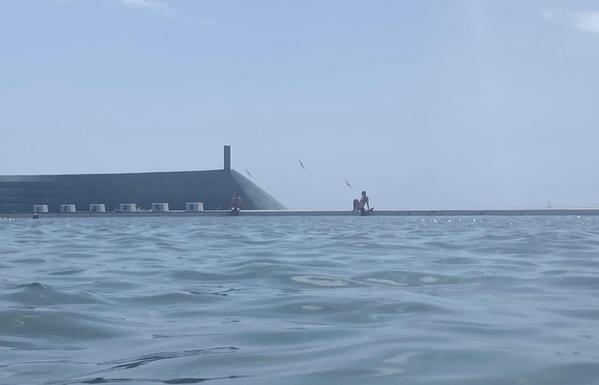 Eastward view from in the ocean baths (taken while swimming) on a mild summer day. Shades of blue: blue-green water, dark turquoise wall, lighter sky. In the distance a swimmer is silhouetted sitting on the edge of the pool talking to a friend in the water whose elbow rests on the wall. Three gulls are flying over, barely more than elongated Ms.