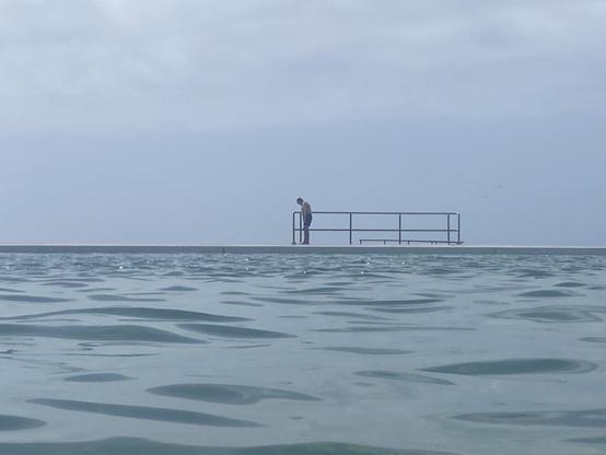 Looking eastwards from within the main pool. A man in bathers is standing in front of the first of three lengths of metal fencing, looking down, perhaps in contemplation. Grey-blue water, paler hazy blur sky. A feeling of quietness.