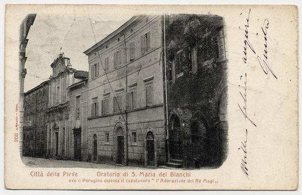The image shows a vintage postcard featuring black and white photography. The building in the center appears to be an old, multi-story structure with classical architectural elements such as pilasters, arched windows, and decorative cornices above ground-level openings. A distinctive feature is a tower on top of another section that extends beyond the main body of the building.

On the left side of the postcard, there's text indicating "Città della Pieve" followed by additional information about "Oratorio di S. Maria dei Bianchi," which translates to "Chapel of St. Mary of the Whites." Additional phrases mention a painting and possibly historical or religious significance associated with this chapel.

The right side has handwritten cursive script, likely indicating ownership or perhaps annotations related to the image's content or history. The top-right corner features another signature that appears more formal than the handwriting on the left. There are visible signs of wear and tear, such as creases and spots, suggesting considerable age.

In summary, this postcard captures a historical chapel in Città della Pieve with handwritten notes likely added by past owners or for descriptive purposes.