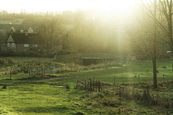 Photo of a green low laying field with a small stream and a bridge.
There are some houses on the left and right is glow out by a low sun.