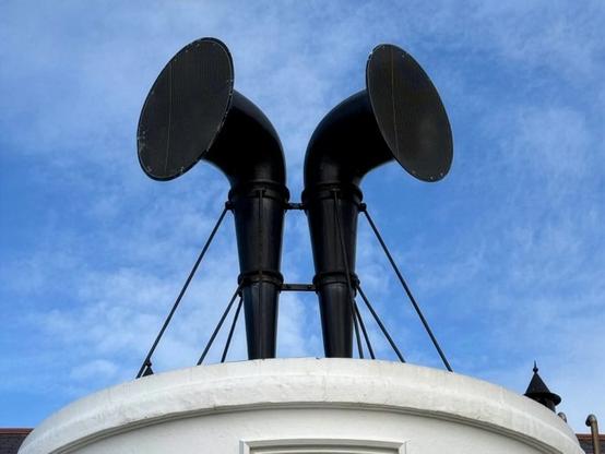 Two large black speakers resembling horns mounted on a white circular structure against a blue sky.