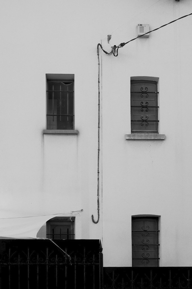 Photo format portrait en noir & blanc d’un mur blanc, avec 4 fenêtres disposée en carrée, celle en bas à gauche à moité cachée par une benne. Un fil électrique noir arrive dans le cadre par le coin supérieur droit, et viens se coller au mur, et descend entre les fenêtres, pour se finir en crochet avant d’entrer dans le mur.