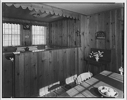 The image depicts a vintage kitchen with wooden paneling on the walls and floor, creating an antique ambiance. A large window allows natural light to flood into the room, highlighting various features such as built-in cupboards, a sink area with a faucet, and decorative elements like wall sconces, mirrors, and flower arrangements.

The dining table in front of the windows is set for two people, adorned with plates, glasses, and bowls. The simplicity of the furnishings and decor suggests practicality over luxury. This setting evokes a sense of history and nostalgia, possibly reflecting life during an earlier era before modern amenities became commonplace.